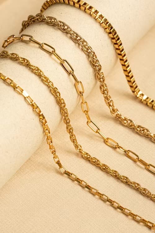 Assorted gold chain necklaces in rope, paperclip, and box-link styles on a cream fabric roll