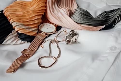Rose-gold mesh watch, chain necklace, and crystal ring arranged on white shirt fabric