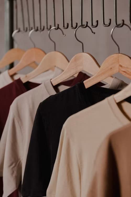 Six folded organic cotton crew-neck t-shirts in red, black, white, navy, burgundy, and yellow on a wooden surface